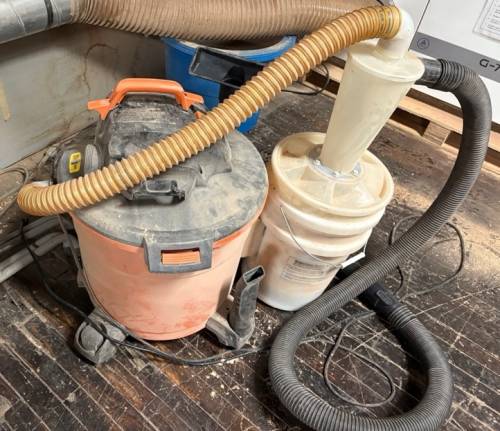 Shop vac on the left, vortex bucket on the right. The tan hose runs from the vac inlet to the top outlet of the vortex. The black hose, on the side of the vortex, goes to the tool. Shop vac on the left, vortex bucket on the right. The tan hose runs from the vac inlet to the top outlet of the vortex. The black hose, on the side of the vortex, goes to the tool.
